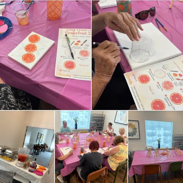 A creative art event featuring participants watercolor painting grapefruit-themed designs. The tables are decorated with pink tablecloths and filled with art supplies. Some participants are actively painting, while others are engaged in conversation. Refreshments are also visible on the tables.