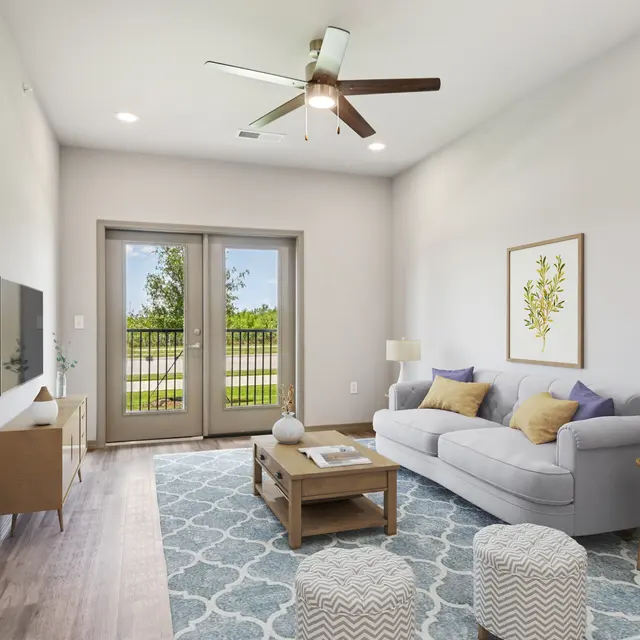 Modern living room featuring a light grey sofa, a coffee table, and decorative poufs on a patterned rug. Large windows provide a view of greenery outside.