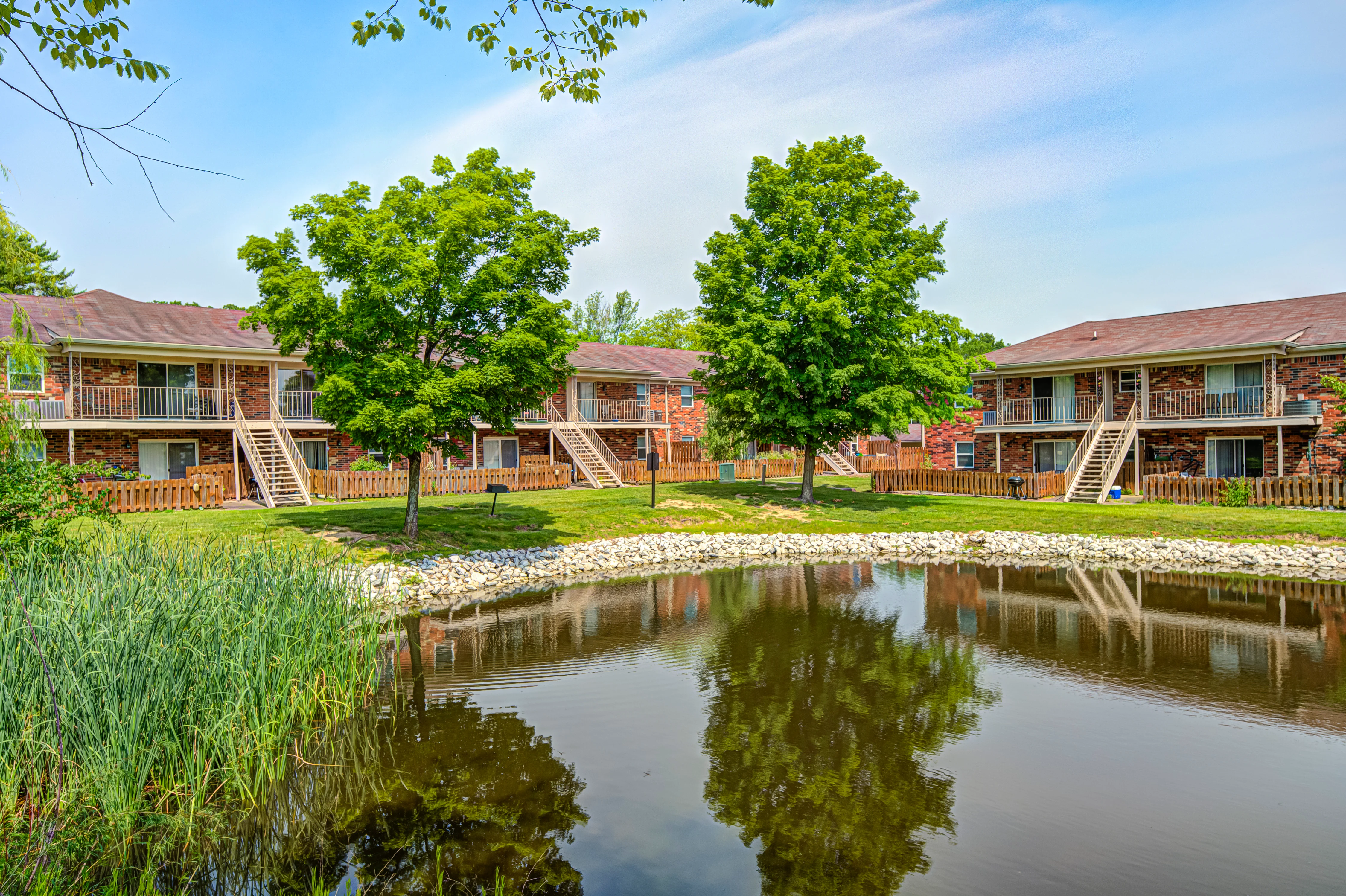Scenic Apartment Complex by a Pond A serene view of a multi-unit apartment complex beside a pond, featuring several trees and manicured grass.