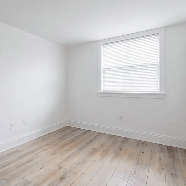 A bright and empty room with white walls and light wooden flooring. There is a window with blinds letting in natural light and a closet with sliding doors on one side.