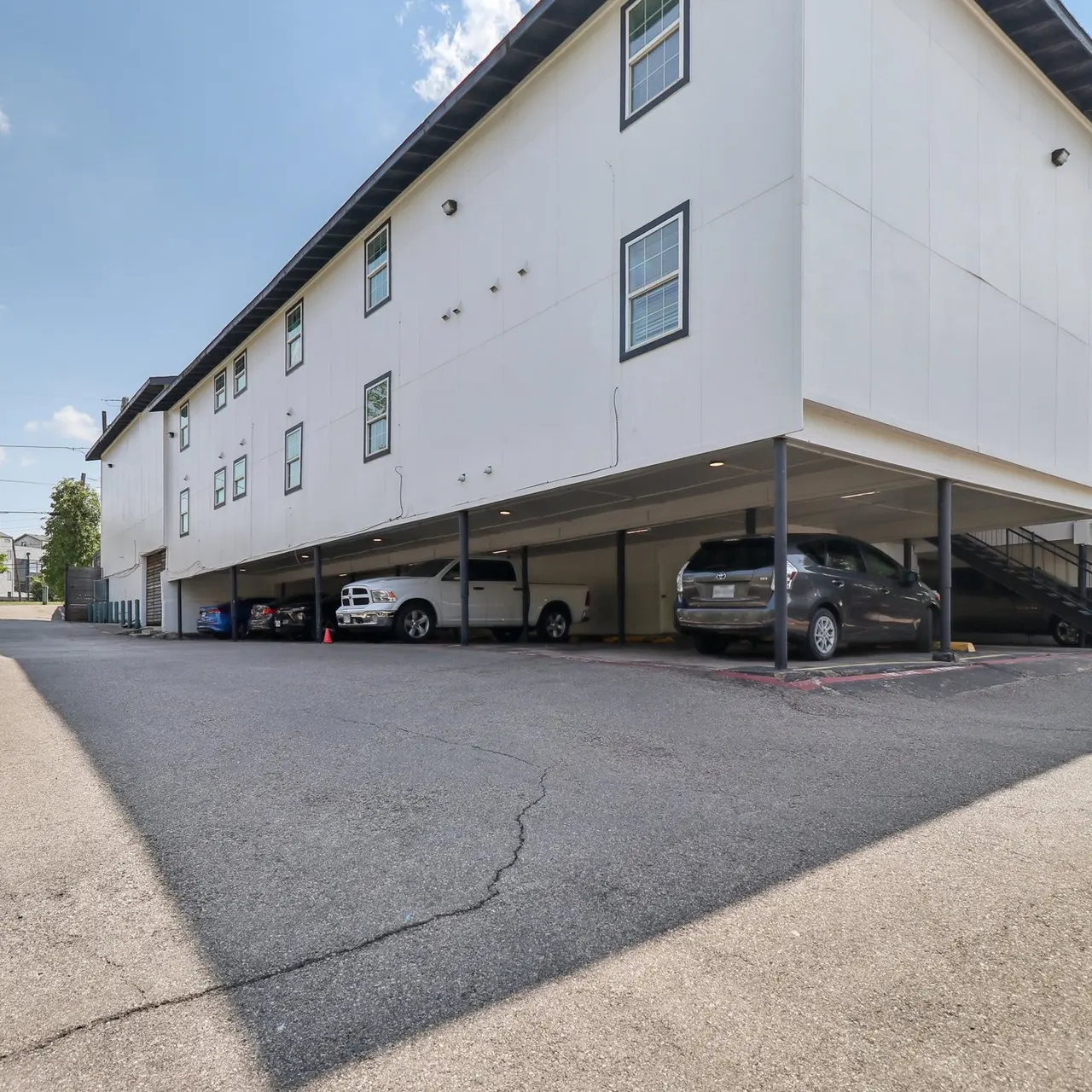 A parking lot with several vehicles parked under a white multi-story building. The building has multiple windows and a staircase visible on the side. The ground is paved with visible markings, and the sky is mostly clear with a few clouds.