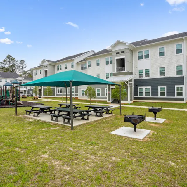 A green space with picnic tables, a playground, and multiple buildings in the background under a clear blue sky.