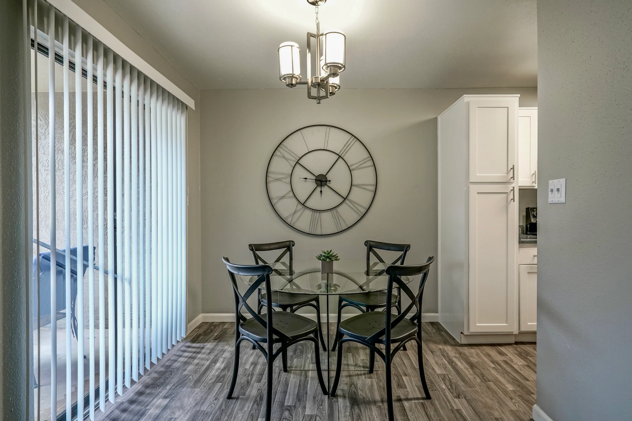 Elegant Dining Area A dining area featuring a circular glass table surrounded by black chairs. A large wall clock with a round design is displayed on the wall. The space is illuminated by a stylish chandelier, and there are sliding glass doors leading outside.