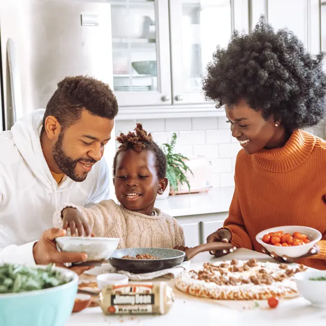 A family cooking in a bright kitchen. A father and mother help their daughter add ingredients to a bowl while smiling and interacting. The kitchen features items like vegetables and fruits on the counter.