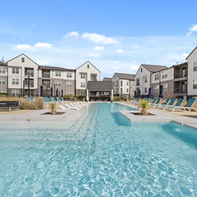 A scenic view of a modern apartment complex featuring a long swimming pool surrounded by lounge chairs and palm trees under a clear sky.
