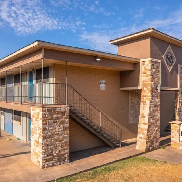 Exterior view of a two-story apartment complex featuring tan walls, stone accents, and a staircase. The image shows parked cars and grass in the foreground, with signs indicating the property name.