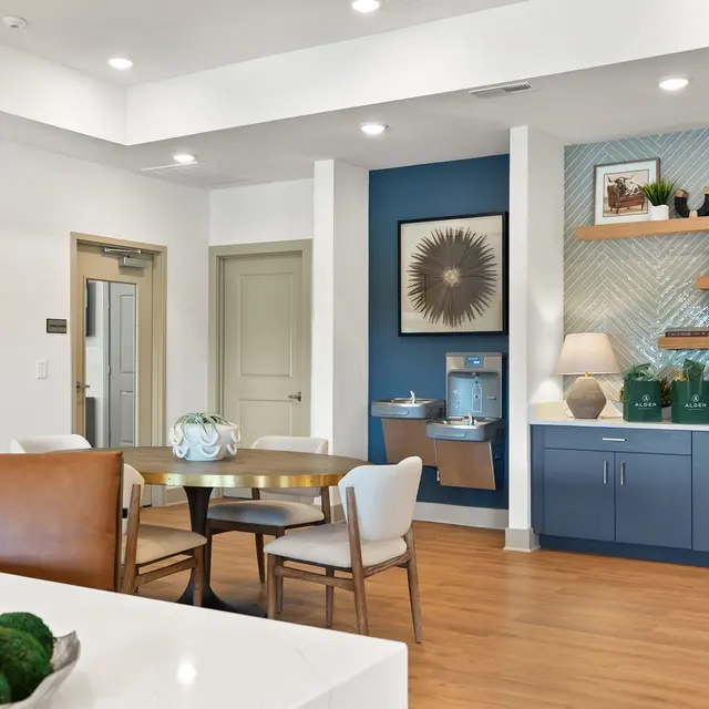 A modern and stylish interior space featuring a dining table with chairs, decorative wall art, a water cooler, and well-styled shelves. The room has hardwood flooring, bright lighting, and a cozy atmosphere.