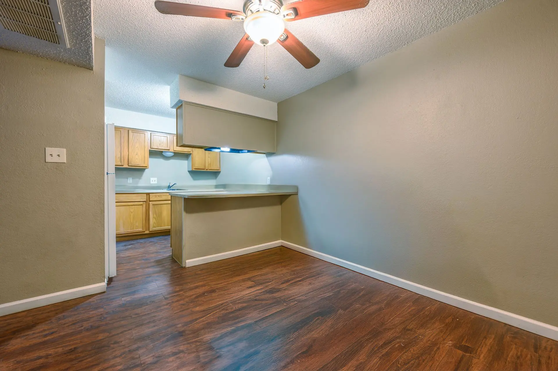 A spacious living area in an apartment with wooden flooring and a ceiling fan. The kitchen is partially visible in the background with wooden cabinets and a light green wall.