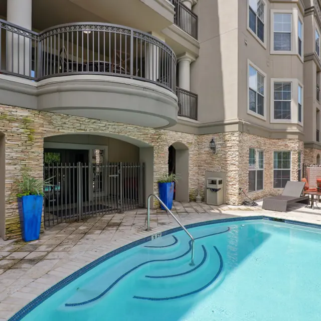 A modern apartment complex featuring a small pool area with a cozy seating space. The pool is surrounded by stone and has a gentle wave design on the bottom. Lounge chairs are placed around the pool, with bright blue flower pots adding a decorative touch near the entrance. The building features balconies above the pool area, with greenery visible in the background.