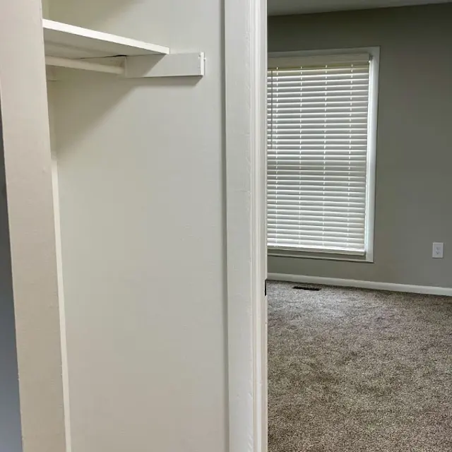A view between two doorways showing a room with a window and carpeted floor, and a small closet space partially visible alongside.
