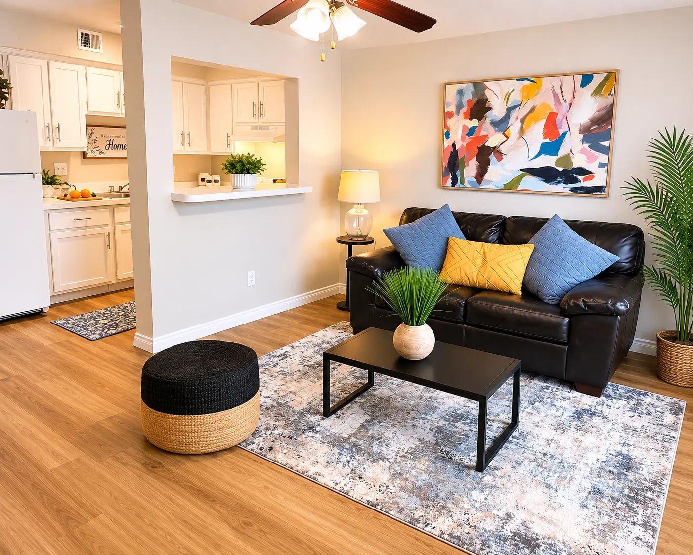 A stylish living room featuring a black leather sofa adorned with two blue pillows and a yellow pillow, a low black coffee table, and a decorative plant. The flooring is wooden, and a colorful abstract painting is mounted on the wall. The open kitchen is visible in the background, with a white refrigerator and a small portion of counter space.