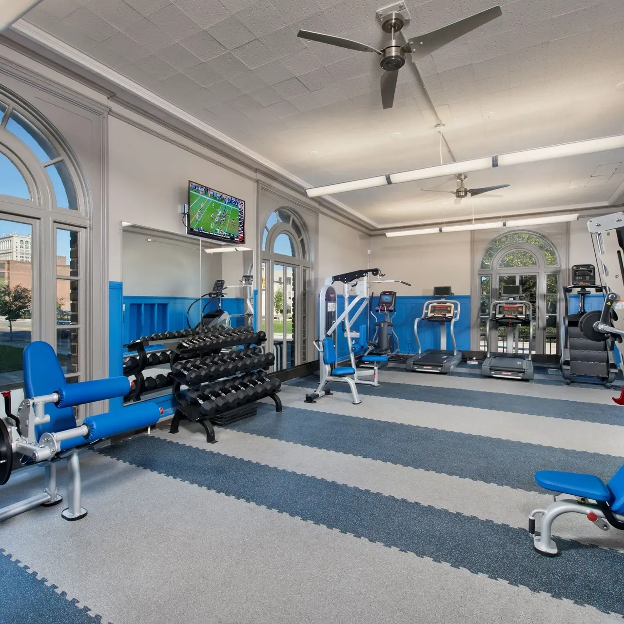 A modern fitness gym featuring various exercise equipment including free weights, stationary bikes, and cardio machines. Large windows provide natural light, and a TV is mounted on the wall.