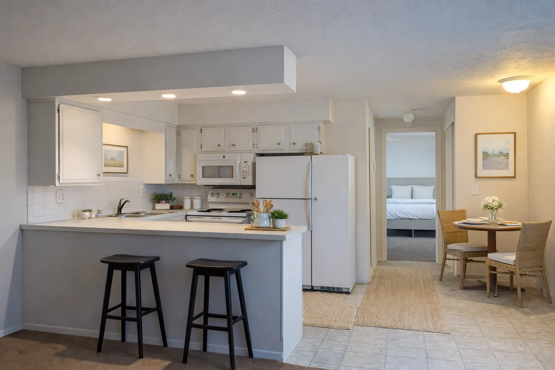 A modern kitchen with a breakfast bar featuring two black stools, white cabinetry, and a refrigerator. The space is well-lit and opens to a dining area with a small table and chairs. A doorway in the background leads to a bedroom.