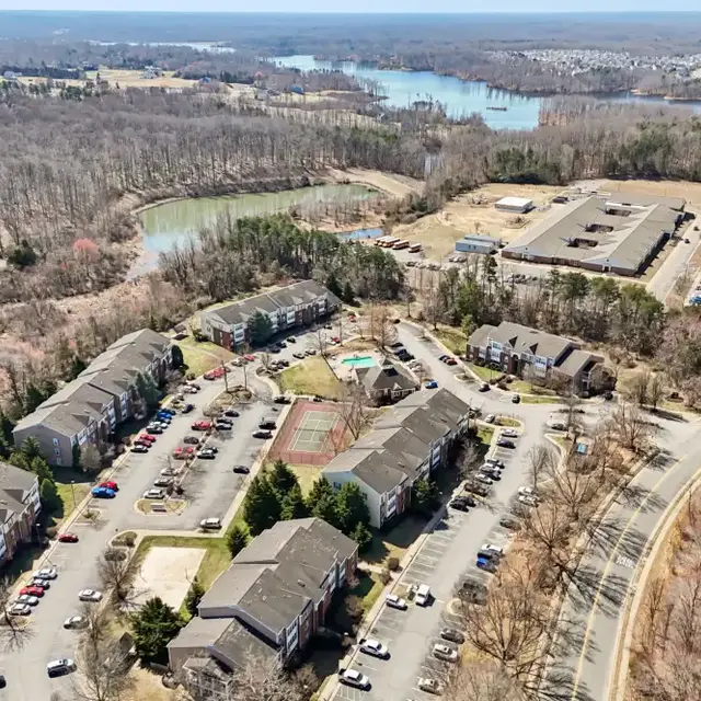 Aerial view of a suburban apartment complex with parking lots, green areas, and a nearby lake in the background.