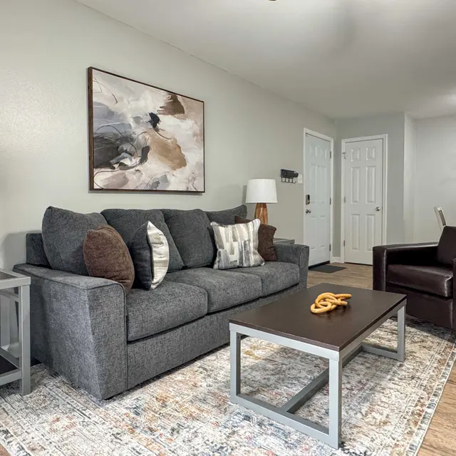 A modern living room featuring a gray sofa with decorative pillows, an armchair, a wooden coffee table with a decorative piece on it, and warm lighting, set against a neutral wall with artwork and a dining area visible in the background.