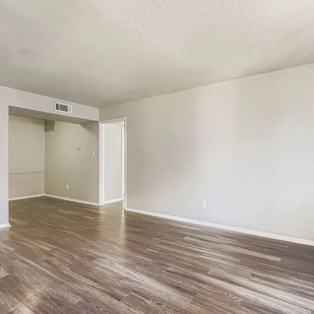 An empty living room with wooden floors and light-colored walls. A doorway leads to another room on the left.
