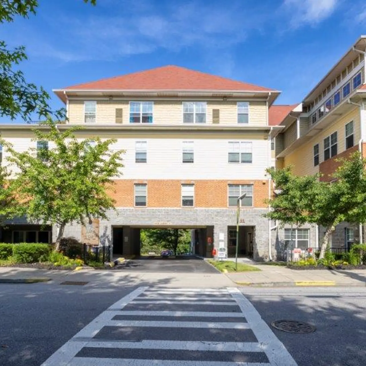 A multi-story apartment building with a red roof and cream-colored walls, surrounded by green trees, under a clear blue sky.