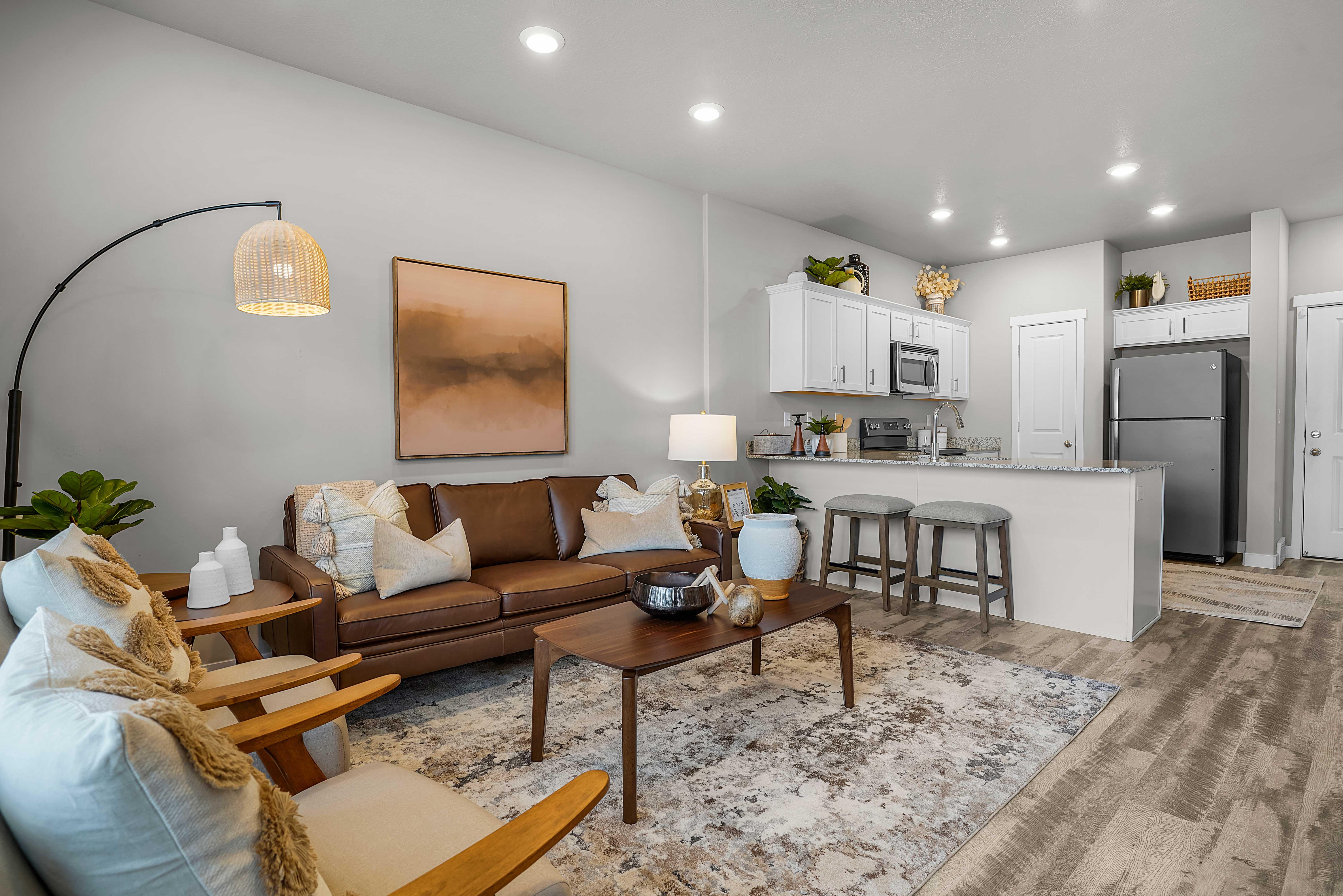 A cozy living room with a brown leather sofa, white armchairs, and a wooden coffee table. The space features light-colored walls, a modern art piece, and a kitchen area with white cabinetry and stainless steel appliances.