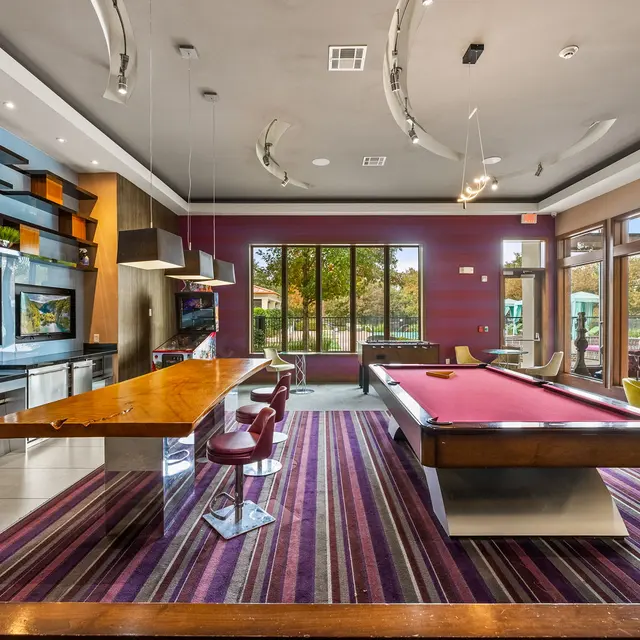 A modern game room featuring a billiards table, dining area with stylish chairs, large windows providing natural light, and a sleek kitchen area. The decor includes a mix of wood and colorful accents on the walls.