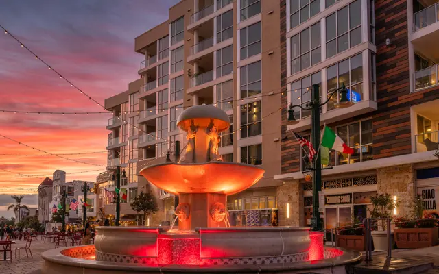 A beautifully lit fountain in a plaza surrounded by modern buildings at sunset. The sky displays vibrant hues of pink and orange, reflecting off the water, while decorative lights are strung overhead.