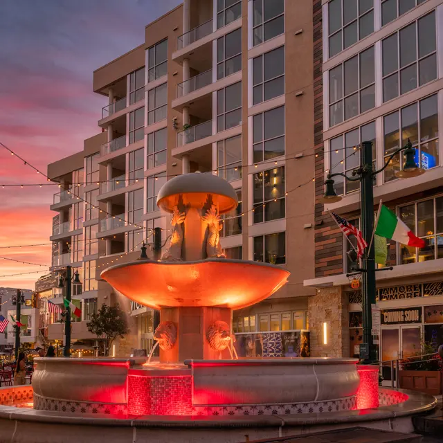 A beautifully lit fountain in a plaza surrounded by modern buildings at sunset. The sky displays vibrant hues of pink and orange, reflecting off the water, while decorative lights are strung overhead.