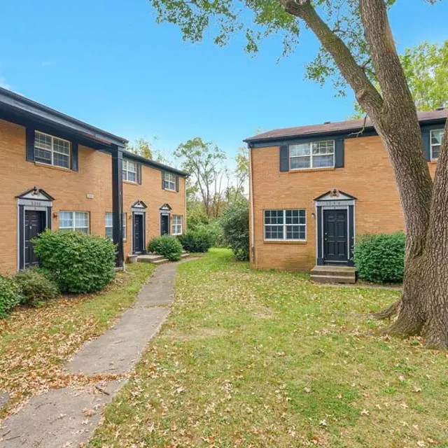 A view of two brown brick apartment buildings in a residential area with green grass and shrubs.