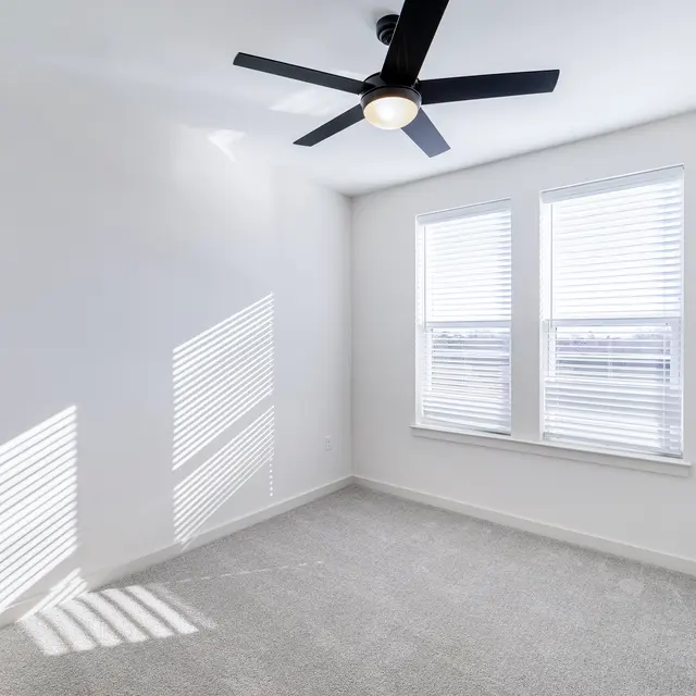 A bright, empty room with light gray carpet and two windows with white blinds. A ceiling fan is mounted on the ceiling, and shadows from the blinds create striped patterns on the wall.