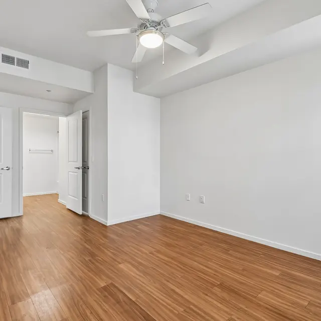 Empty Room with Ceiling Fan A spacious empty room with light-colored wooden flooring, a ceiling fan, and white walls. Two doors are visible, one leading to a closet, and the other opening into another space. The room is well-lit with natural light coming from an unseen window.