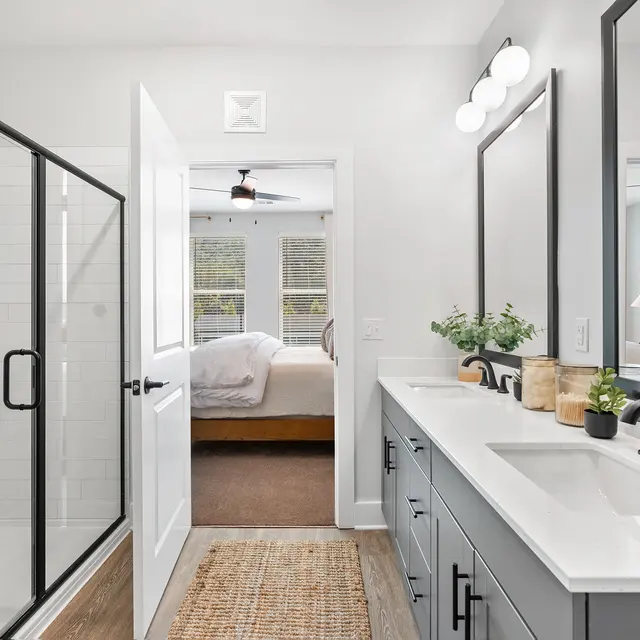 A modern bathroom featuring a glass shower, double sink vanity with dark cabinetry, and a doorway leading to a bedroom with a large bed and large window, illuminating the space with natural light.