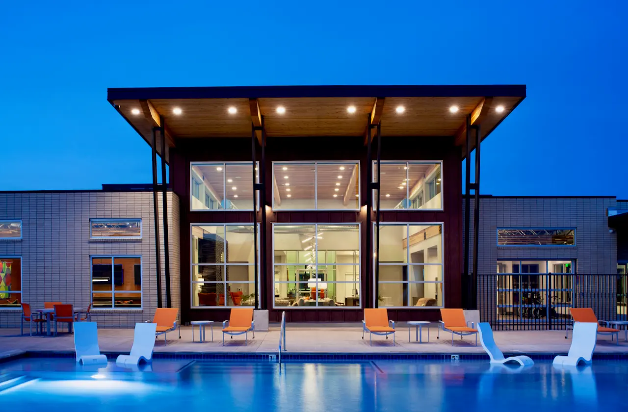 A modern poolside clubhouse at dusk, featuring large glass windows and a contemporary design structure. The pool is illuminated, with lounge chairs around it and a blue sky above.