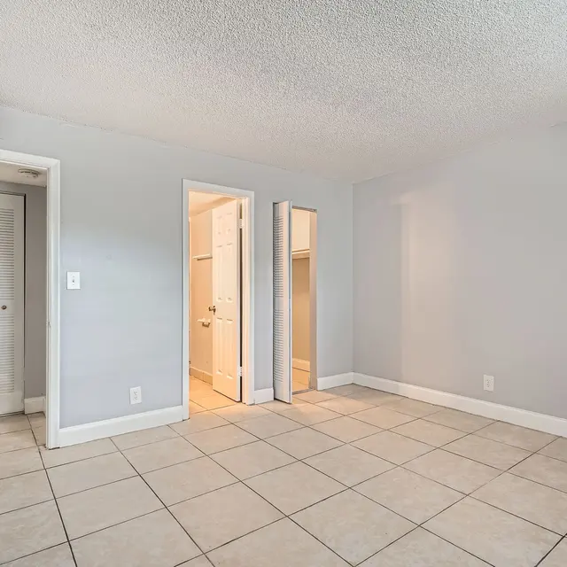 A spacious, empty room with light gray walls and tiled flooring. There are two doors visible, one leading to a closet and another to a bathroom. The room has a neutral aesthetic with natural light coming from the entrance.