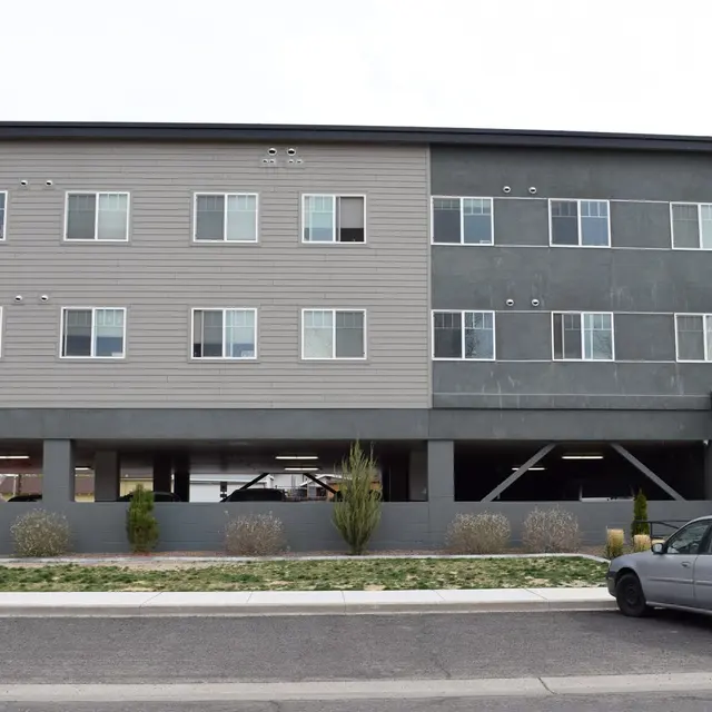 Contemporary Apartment Building A modern three-story apartment building with a mix of beige and grey siding, featuring multiple windows and a flagpole with an American flag in front.