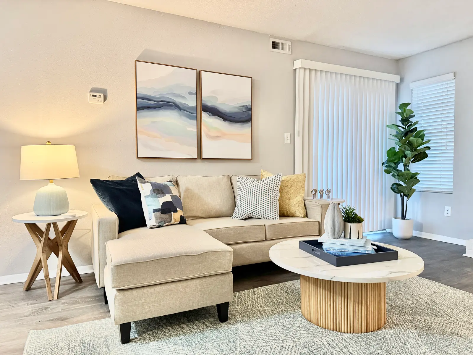A modern living room featuring a beige sectional couch with colorful throw pillows, a round coffee table, a lamp, and large artwork on the wall. Natural light streams in through the blinds.