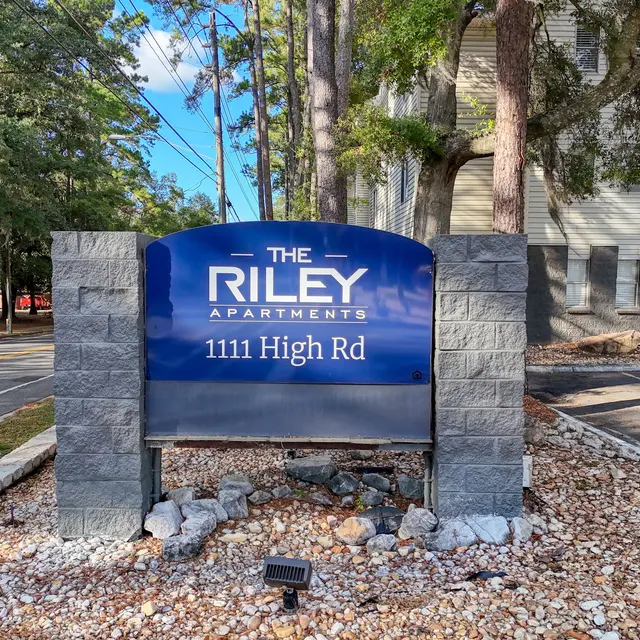 Sign for The Riley Apartments located at 1111 High Rd, surrounded by trees and pebbled landscaping.