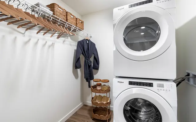 Modern Laundry Room A modern laundry room featuring a stacked washer and dryer, a blue bathrobe hanging on a rack, and several baskets for storage. The walls are painted white and the flooring is wooden.