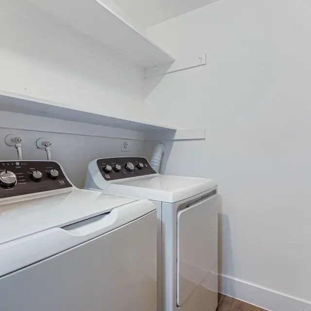 A laundry room featuring a washing machine and dryer next to each other, with white shelves on the wall for storage, and wooden flooring.