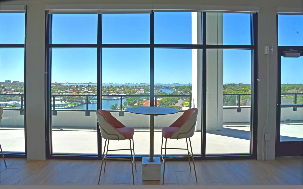 A modern space with a round table and chairs facing large windows, showcasing a scenic view of a river and houses outside.
