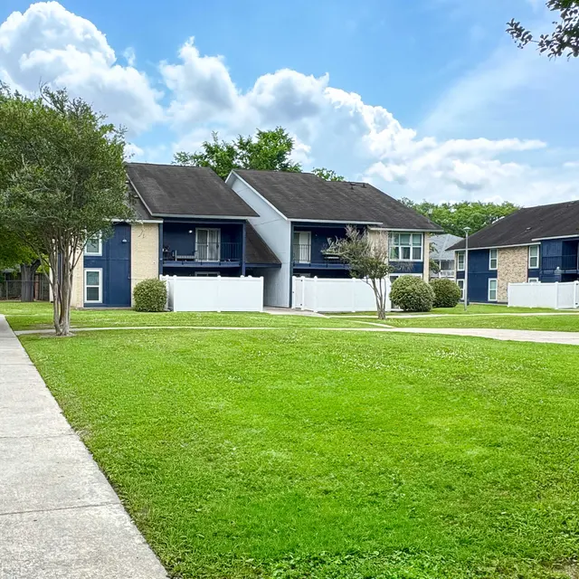 View of an apartment complex with multiple buildings, green grass, and a clear blue sky with clouds.