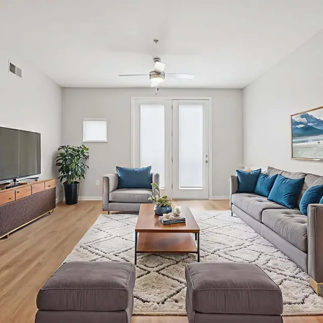 Modern Living Room Interior Spacious and modern living room featuring a grey sectional sofa with blue cushions, a wooden coffee table, a flat-screen television, and large windows allowing natural light. The room has light-colored walls and a decorative painting on the wall.
