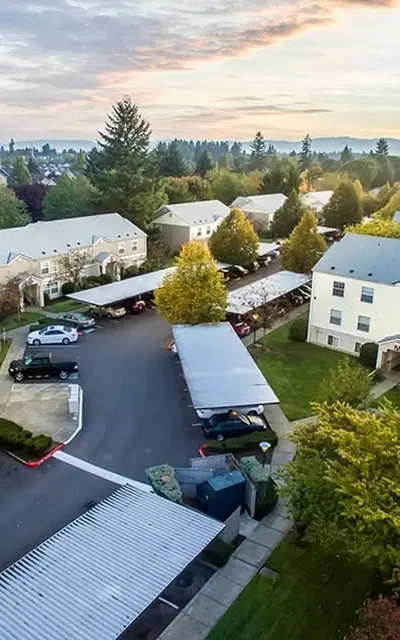 Aerial view of a residential neighborhood with several buildings, parking areas, trees, and a scenic backdrop of hills and a sunset sky.