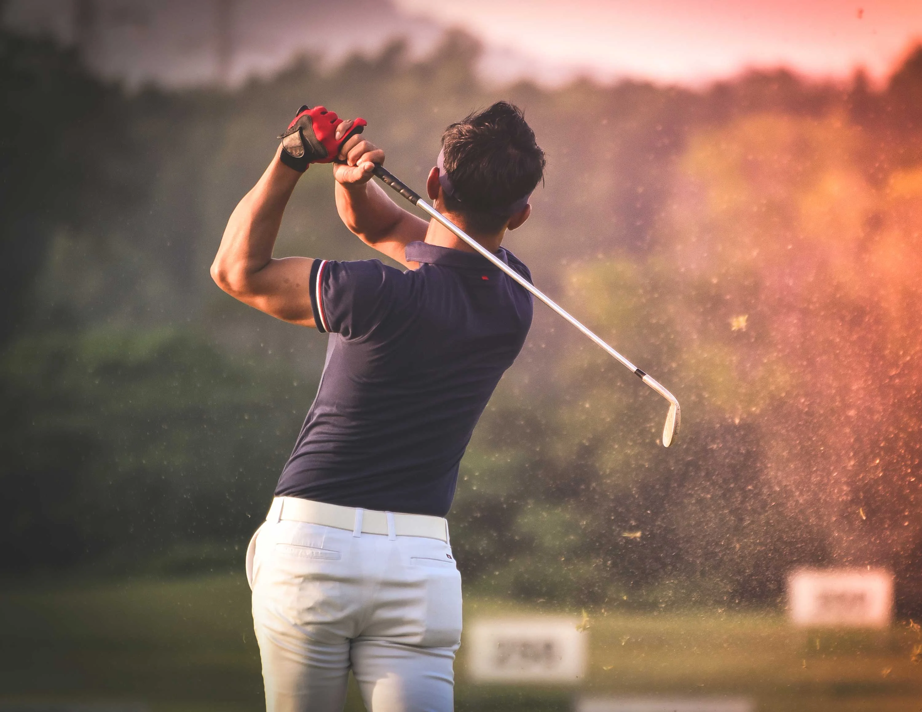 The Hideaway A golfer in a dark shirt and white pants is swinging a club on a golf course with trees in the background, capturing a moment of action with dust flying up.