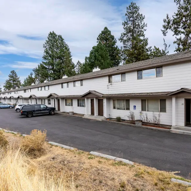 A row of two-story apartment buildings with a parking lot in front, surrounded by trees and grass.