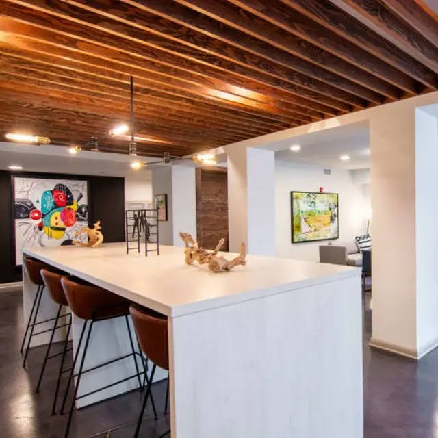 A modern interior space featuring a long bar-like kitchen counter with brown stools, stylish artwork on the walls, and warm wooden ceiling beams.