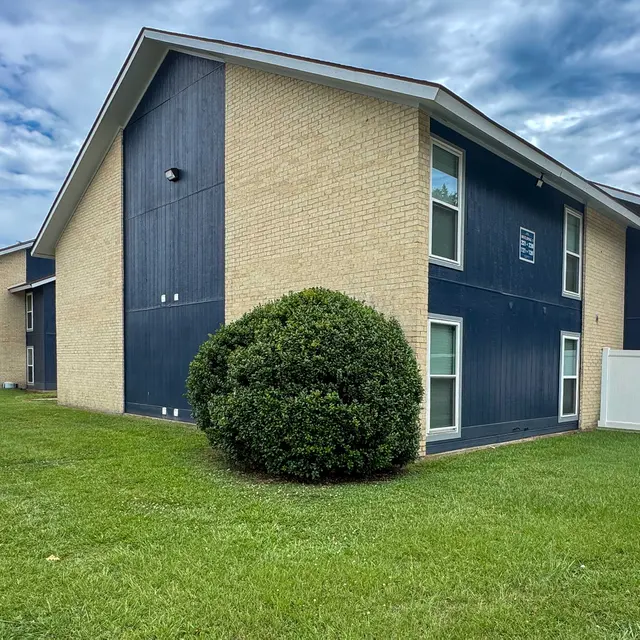 Exterior view of a residential apartment building with a dark blue side and light brick facade, surrounded by grass and some clouds in the sky.