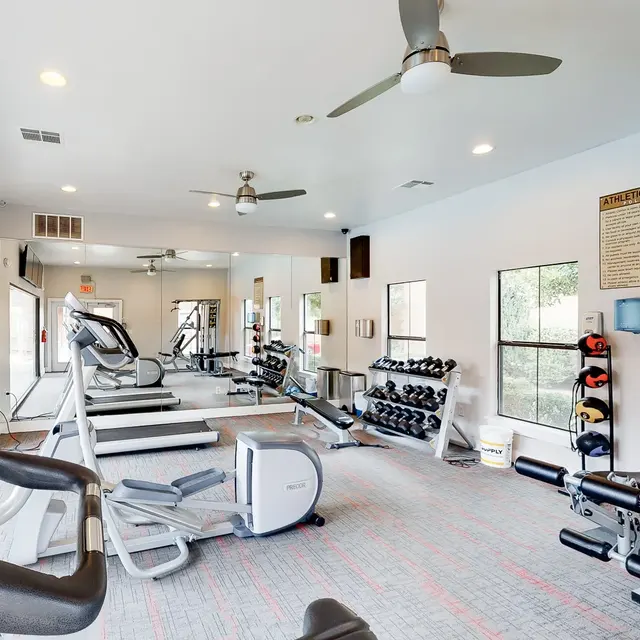 A spacious gym interior with exercise equipment including treadmills, weights, and benches, bright overhead lights, and large windows letting in natural light.