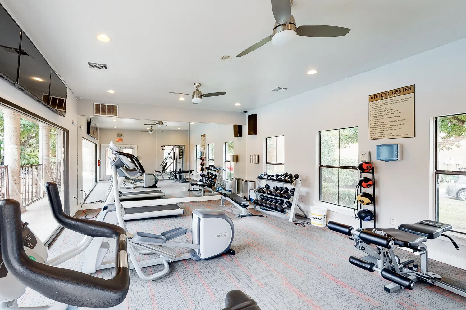 A spacious gym interior with exercise equipment including treadmills, weights, and benches, bright overhead lights, and large windows letting in natural light.