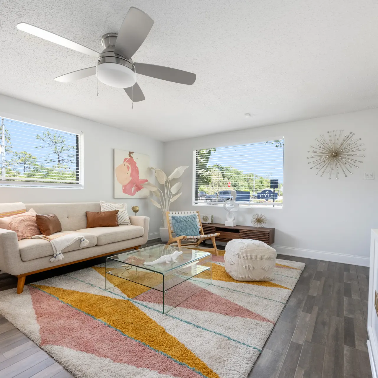 A bright and modern living room featuring a light-colored sofa, decorative pillows, a glass coffee table, and a patterned area rug with pastel colors. A large window provides natural light, and a decorative floor lamp stands in the corner.