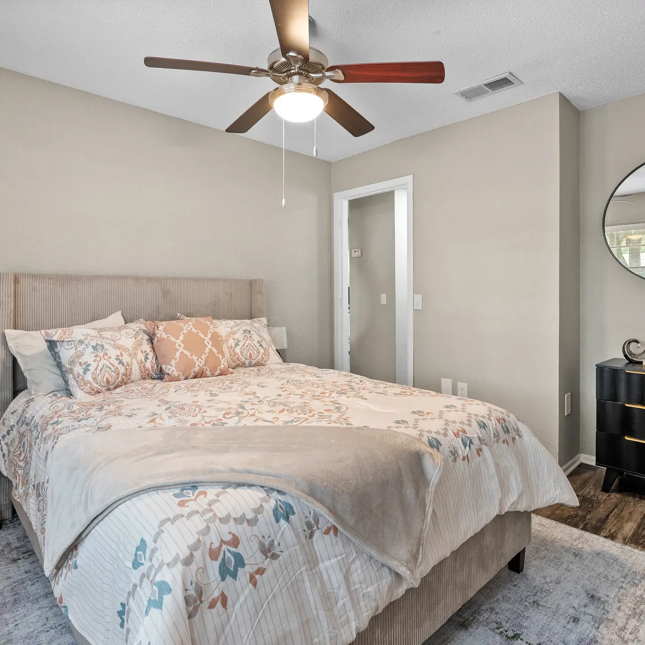 A well-decorated modern bedroom featuring a queen-sized bed with decorative pillows, a stylish nightstand, and a black dresser. A mirror hangs on the wall, reflecting natural light from a window.