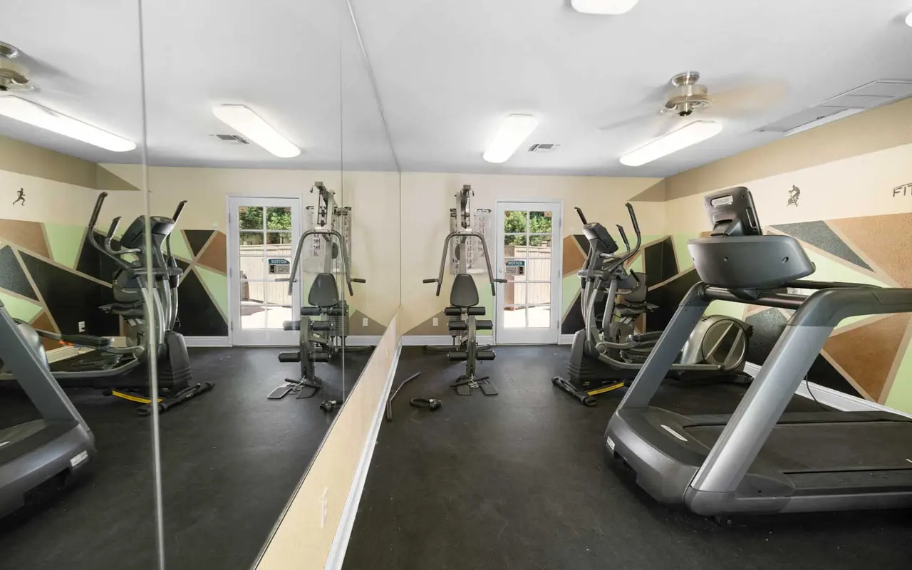 A modern gym interior featuring exercise equipment, including treadmills and a multi-gym machine. Large mirrors reflect the equipment, and the walls have a geometric design. Windows provide natural light.