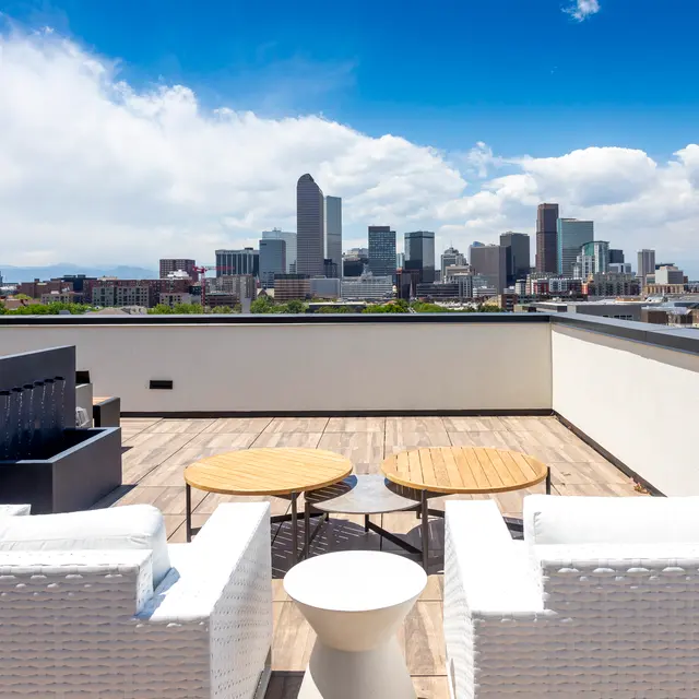 A rooftop terrace with modern furniture overlooks a city skyline under a bright blue sky with scattered clouds.